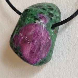 Può includere: Un ciondolo di pietra preziosa lucidata con un cordino nero. La pietra è una miscela di verde e rosa intenso, con una zona rosa prominente. Il ciondolo è liscio e arrotondato, con un foro per il cordino.