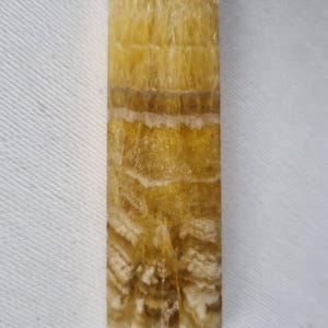 Peut inclure: Une pointe de cristal jaune et marron avec un corps rectangulaire et un sommet pointu. Le cristal a une apparence en couches avec des bandes de couleur et une surface texturée. La base du cristal a une légère teinte violette.