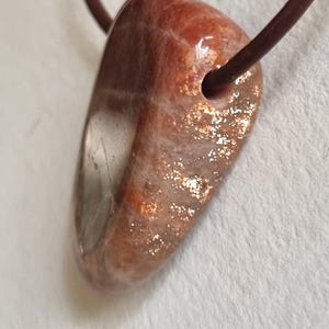 Op de afbeelding: Een gepolijste, driehoekige zonnesteen hanger met een roodbruine tint en metallic gouden insluitingen, hangend aan een dun, donkerbruin leren koord. De steen heeft een glad, glanzend oppervlak met zichtbare interne glinsteringen en een boorgat.