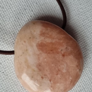 Puede incluir: Un collar con colgante liso de forma ovalada. El colgante es de color melocotón claro con marcas melocotón y marrón más oscuras. Un cordón marrón oscuro está unido al colgante. El collar se muestra sobre una superficie blanca texturizada.