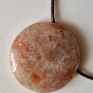 Può includere: Un ciondolo rotondo in pietra solare con un cordino in pelle marrone. Il ciondolo è di colore beige chiaro con macchie arancioni e dorate. La pietra ha una finitura lucida e lucente. Il cordino è sottile e sembra essere annodato.