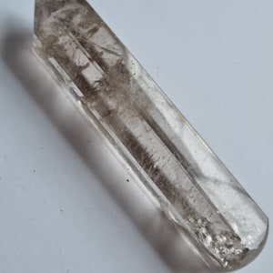Può includere: Un cristallo di quarzo trasparente con una tonalità fumosa marrone chiaro. Il cristallo è rettangolare con una parte superiore appuntita e una parte inferiore arrotondata. La superficie presenta linee e imperfezioni naturali. È una gemma naturale.