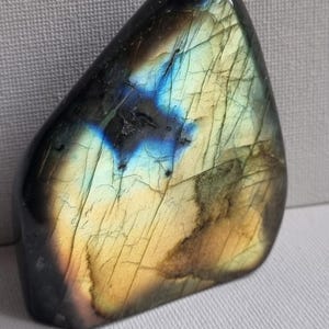 Könnte beinhalten: Ein polierter Labradorit-Edelstein in dreieckiger Form. Er zeigt ein schillerndes Farbspiel mit Blau-, Grün- und Gelbtönen. Die Oberfläche weist natürliche Risse und Variationen auf. Ein natürlicher, dekorativer Gegenstand.