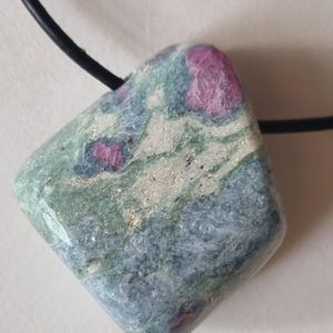 Könnte beinhalten: Ein quadratischer Anhänger aus Naturstein mit einer Mischung aus grünen, blauen und weißen Farbtönen mit rosa Flecken. Der Anhänger ist an einer schwarzen Kordel befestigt, wodurch eine Halskette entsteht. Der Stein hat eine polierte Oberfläche.