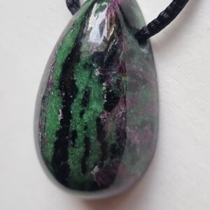 Può includere: Un ciondolo a forma di goccia con un cordino nero. Il ciondolo è una pietra lucidata con un mix di colori verdi, neri e viola. La pietra ha una finitura lucida ed è probabilmente una pietra preziosa.
