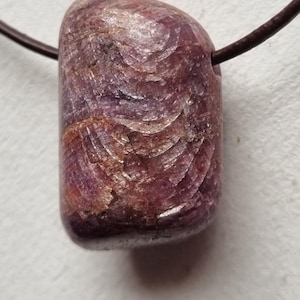 Peut inclure: Un pendentif cylindrique en pierre précieuse polie avec un cordon marron foncé. La pierre est un mélange de teintes violettes profondes et brunes, avec un motif texturé et tourbillonnant sur sa surface. Le pendentif est probablement un collier.