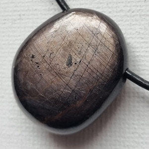 Può includere: Una collana con cordoncino in pelle nera con un ciondolo ovale di colore grigio scuro. Il ciondolo ha una superficie testurizzata con un motivo di linee sottili.