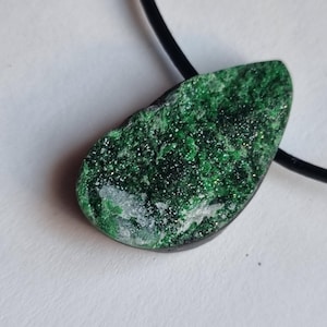 Op de afbeelding: Een groene edelsteen hanger aan een zwart koord. De hanger heeft een druppelvorm met een ruw, getextureerd oppervlak. De steen is diepgroen met sprankelende insluitsels, gezet in een donker frame.