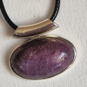 Op de afbeelding: Een hangerketting met een grote, ovaalvormige paarse steen in een gepolijste, zilverkleurige metalen zetting. De hanger zit aan een glad, zwart koord dat door een gebogen, zilverkleurige metalen hangerhouder aan de bovenkant loopt.