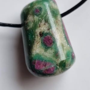 Può includere: Un ciondolo di pietra preziosa lucidata con base verde e inclusioni rosse. La pietra è a forma di botte ed è infilata su un cordino nero. Il ciondolo è lungo circa 2,5 cm.