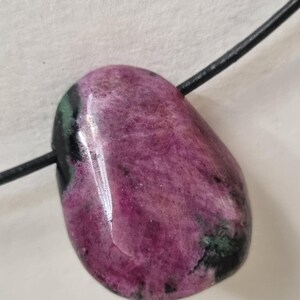 Può includere: Un ciondolo di pietra preziosa lucidata, di forma ovale. La pietra è di un colore magenta intenso con striature rosa più chiare e inclusioni verde scuro. È attaccato un cordino nero, che suggerisce una collana.