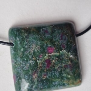 Può includere: Un ciondolo quadrato con una pietra screziata verde e rossa. La pietra ha una superficie lucida ed è attaccata a un cordino nero. Il ciondolo è una pietra preziosa naturale, forse giada o diaspro.