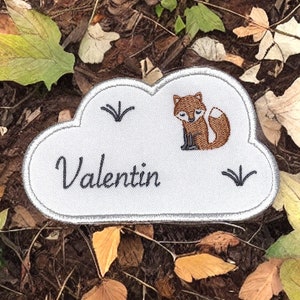 以下が含まれることがあります： 白い雲の形をした刺繍パッチで、名前は「Valentin」と、茶色のキツネのイラストが描かれています。パッチはグレーの縁取りと緑の草の刺繍が施されています。