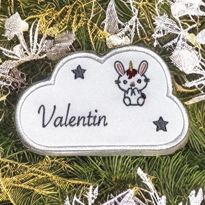 以下が含まれることがあります： 「Valentin」という名前と小さなユニコーンのイラストが描かれた、白い雲の形をした刺繍パッチ。パッチには小さな星が2つ付いています。