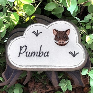 Peut inclure: Un patch brodé en forme de nuage blanc avec le nom "Pumba" et une image de phacochère en dessin animé. Le patch est fixé sur un support en bois avec des pieds bruns.