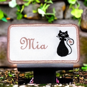 Puede incluir: Parche blanco bordado con un borde rosa. El parche presenta el nombre "Mia" y un gato negro con un corazón rosa en la cola.