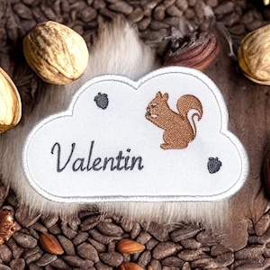 以下が含まれることがあります： 白い雲の形をした刺繍パッチ。茶色のリスがナッツをくわえ、黒い筆記体で「Valentin」と書いてあり、小さな茶色のドングリが2つ付いています。パッチは茶色のナッツで囲まれています。
