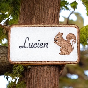 May include: A white rectangular patch with a gold border, featuring the name "Lucien" in cursive script and a brown embroidered squirrel. The patch is attached to a tree trunk.