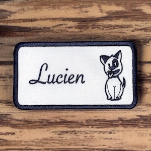 May include: White rectangular embroidered patch with a black border. The patch has the name "Lucien" in cursive script and a cartoon dog illustration.