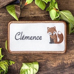 May include: A white embroidered patch with a brown fox design and the name "Clémence" in black cursive lettering. The patch has a brown border and is on a wooden background.