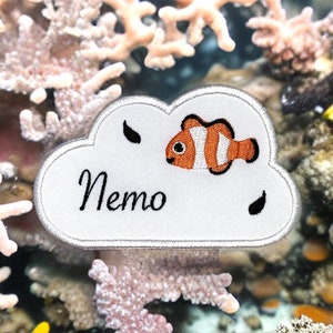 Può includere: Toppa ricamata a forma di nuvola bianca con un pesce pagliaccio arancione e bianco disegnato, la parola "Nemo" e due forme nere a forma di lacrima. La toppa è su uno sfondo di corallo rosa e bianco.