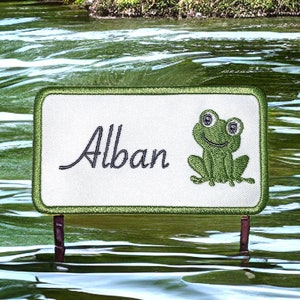 May include: A green frog with a white background and the name "Alban" embroidered in black on a green rectangular patch.