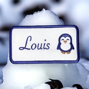 May include: Blue and white embroidered patch with a penguin and the name "Louis" on a white background.