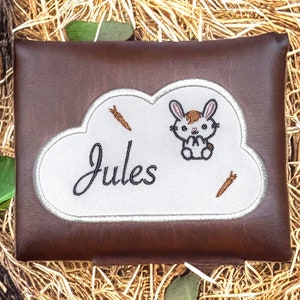 以下が含まれることがあります： 白い雲の形をしたパッチが付いた、茶色の合皮製のジッパー付きポーチ。パッチには、ニンジンを持ったウサギと、筆記体で「ジュール」という名前が刺繍されています。