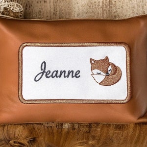 Peut inclure: Un coussin en cuir marron avec un patch brodé blanc représentant un renard endormi et le nom "Jeanne".