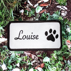 Pode incluir: Remendo retangular branco com texto bordado em preto "Louise" e uma pegada de pata preta. O remendo está em um leito de grama verde e terra marrom.