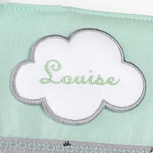 Può includere: Un articolo in tessuto verde menta con una toppa bianca a forma di nuvola ricamata con il nome "Louise" in corsivo verde. La nuvola ha un bordo grigio. Un tessuto a fantasia grigio e bianco è visibile in basso.