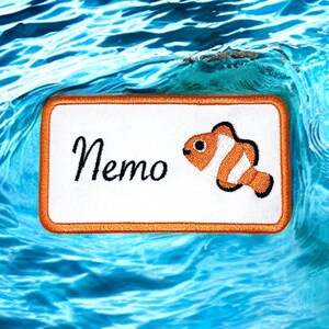 Peut inclure: Patch brodé orange et blanc avec un poisson-clown de dessin animé et le mot "Nemo" sur un fond blanc. Le patch est sur un fond d'eau bleu.