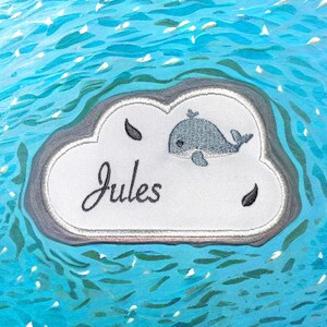 Puede incluir: Un parche blanco en forma de nube con el nombre "Jules" bordado en cursiva negra. Una pequeña ballena azul está bordada encima del nombre. El parche está sobre un fondo azul y blanco.