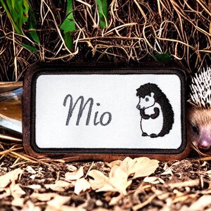 May include: A white embroidered patch with a black hedgehog design and the name "Mio" in gray cursive lettering. The patch is surrounded by a brown border and is lying on a bed of brown leaves.