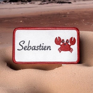 Peut inclure: Patch brodé rouge et blanc avec le nom "Sébastien" et une illustration de crabe rouge. Le patch est sur une plage de sable avec le texte "Le Héron Brodeur" en dessous.