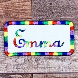 May include: A white rectangular embroidered patch with a rainbow border. The name "Emma" is embroidered in colorful cursive lettering.