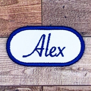 Peut inclure: Écusson ovale blanc avec une bordure bleue. Le nom "Alex" est brodé en lettres cursives bleues.