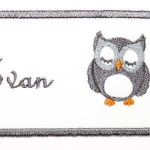 May include: A white embroidered iron-on name tag with a grey owl design and the name "Evan" in cursive script.