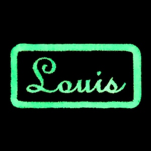 May include: A green glow-in-the-dark rectangular patch with the word "Louis" written in a cursive font.