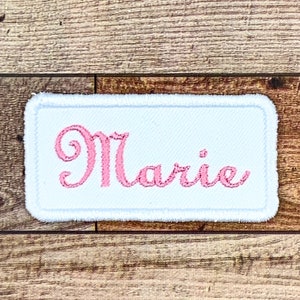 May include: White rectangular fabric patch with pink embroidered lettering that reads "Marie".