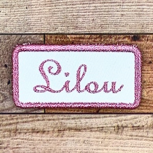May include: A white rectangular patch with a pink border and the name "Lilou" embroidered in pink cursive lettering.