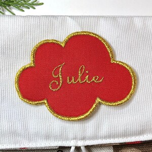 以下が含まれることがあります： 白いストッキングに、赤い雲の形をしたタグが付いています。タグには金色の文字で「Julie」と書かれています。ストッキングには、赤いアクセントのついた茶色のフクロウとベルが付いています。