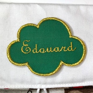 以下が含まれることがあります： 「エドゥアール」と金糸で刺繍された、緑と金の雲の形をした靴下タグです。