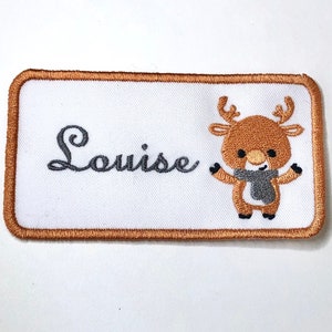 Puede incluir: Un parche rectangular blanco bordado con un borde marrón. El parche presenta el nombre "Louise" en letras cursivas grises y un reno de dibujos animados con una bufanda gris.