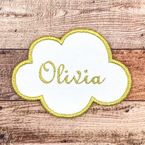 Può includere: Un cerotto bianco a forma di nuvola con un contorno dorato e il nome "Olivia" ricamato in filo dorato.