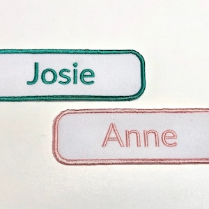 Peut inclure: Deux patchs rectangulaires en tissu blanc avec des noms brodés. Le patch du haut est vert et dit "Josie". Le patch du bas est rose et dit "Anne".