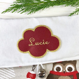 以下が含まれることがあります： 赤い雲の形をした名前タグに金色の文字で「Lucie」と書かれた白いクリスマスストッキング。ストッキングには、赤いスカーフをした茶色のフクロウのデザインとベルが付いています。