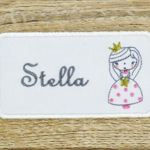 Peut inclure: Étiquette de nom en tissu blanc avec un nom brodé en gris "Stella" et une princesse brodée en rose et blanc avec une couronne dorée.