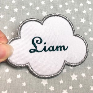 以下が含まれることがあります： 銀色の刺繍の縁取りが施された白い雲の形をした布のパッチ。名前の「Liam」は濃い緑色の糸で刺繍されています。