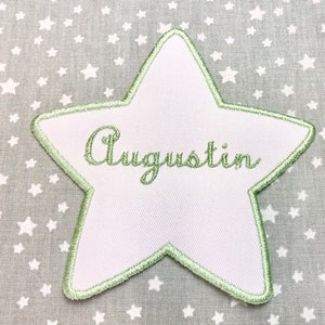 Può includere: Una toppa a forma di stella bianca con un contorno verde. Il nome "Augustin" è ricamato in lettere corsive verdi sulla toppa.